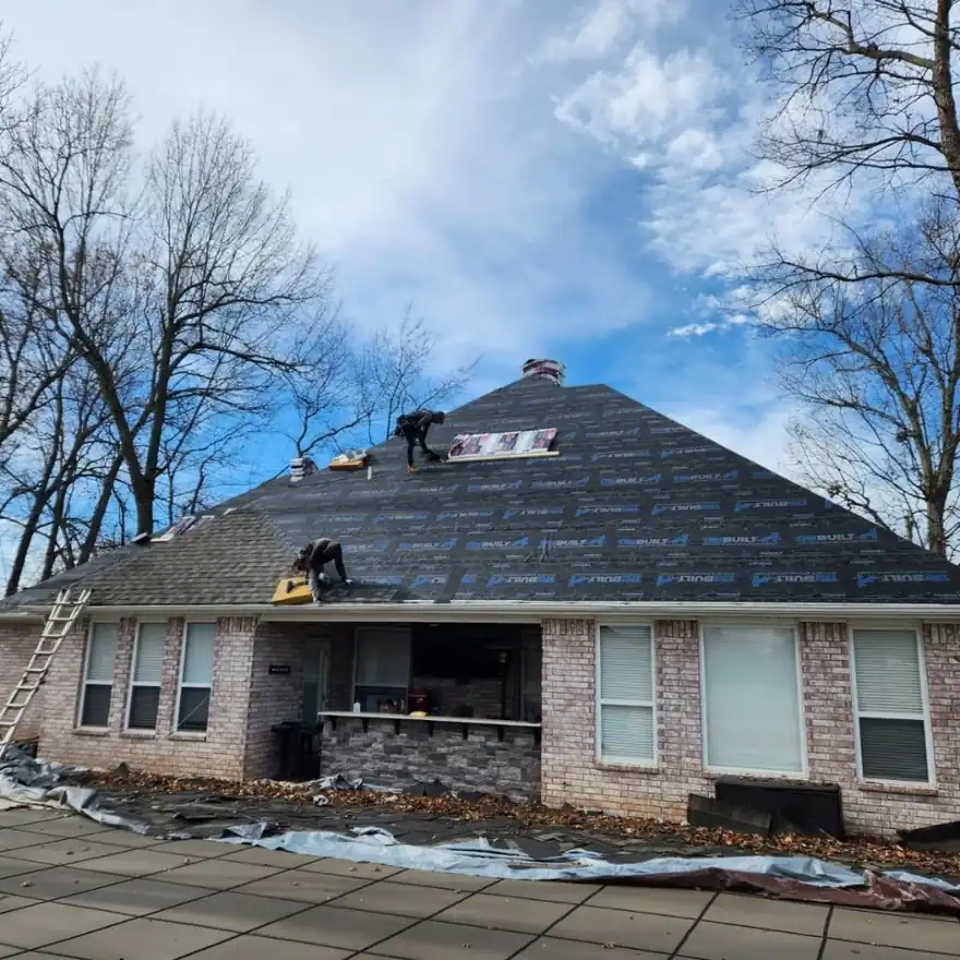 Elite Home Solutions roofers laying synthetic underlayment on a brick ranch's hip roof, ladder at the eave and tarps protecting the patio